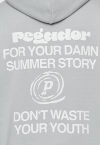 Sudadera gris con texto blanco: "pegador PARA TU MALDITA HISTORIA DE VERANO NO DES APROVECHES TU JUVENTUD," que presenta un logo circular con un gráfico de 'p'.