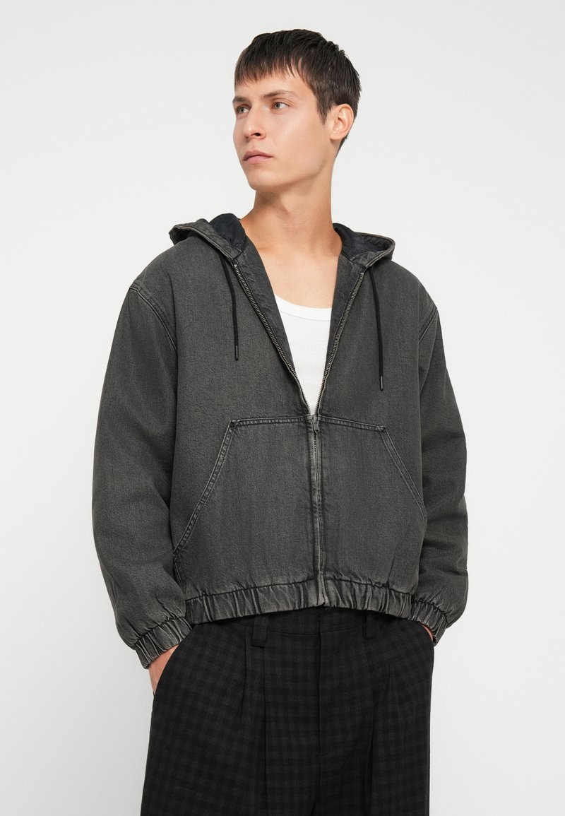 Chaqueta de color gris oscuro con cremallera y capucha, dobladillo elástico y bolsillos frontales. Llevada sobre una camiseta blanca y combinada con pantalones oscuros con estampado.