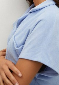 Košilové tričko s krátkým rukávem, světle modré barvy, vyrobené z bavlny, s kapsou a volnějším střihem, s klasickým límcem a měkkou texturou.