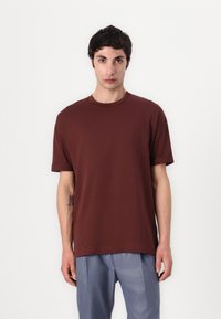 Bruine, korte mouwen t-shirt met een ronde halslijn, gemaakt van katoen. Heeft een relaxte pasvorm en opgerolde mouwen, in combinatie met lichtblauwe broeken.