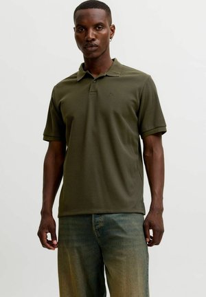 Mann trägt olivgrünes kurzärmeliges Polohemd und blaue Jeans und steht vor einem schlichten hellen Hintergrund.