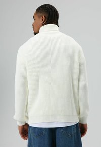 Man met gevlochten haar, gekleed in een witte geribbelde coltrui en een blauwe spijkerbroek, met zijn rug naar de camera tegen een effen grijze achtergrond.