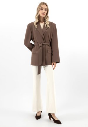 Blonde vrouw met een bruine blazer met riem, witte broek met wijde pijpen en bruine hakken, staand tegen een effen witte achtergrond.