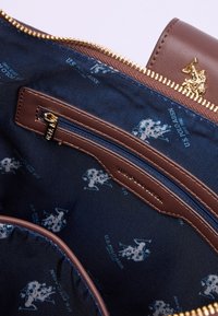 Wnętrze brązowej skórzanej torebki z granatową tkaniną wykończeniową, zdobione powtarzającym się jasnoniebieskim wzorem logo oraz z wewnętrzną kieszenią na zamek.