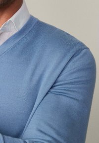 Blauer Strickpullover mit V-Ausschnitt, der eine glatte Textur und eine enganliegende Passform aufweist, über einem weißen Hemd mit Kragen getragen.