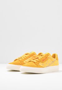 Gula mockaskor med snörningsdesign, med pastellblå accenter och en vit gummi sula. Varumärke på sidan och tungan.