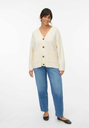 Light cream cardigan with a V-neck, five buttons, ribbed cuffs, and two front pockets paired with blue straight-leg jeans and black flats.