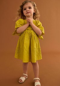 Gelbes besticktes Kleid mit kurzen Ärmeln, das ausgeschnittene Muster aufweist. Kombiniert mit cremefarbenen Sandalen und einer Haarspange, vor einem braunen Hintergrund.