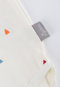 Cremefarbener Stoff mit kleinen blauen, roten und orangefarbenen Dreiecks-Mustern. Beinhaltet ein graues Stoffetikett mit weißem Text.
