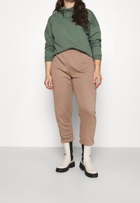 Sudadera con capucha verde con cordón, pantalones deportivos beige ajustados en los tobillos y botas crema hasta el tobillo con detalles en negro y suela gruesa. Texturas de tela suaves.