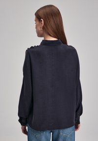 Ciemnoniebieska bluzka z długimi rękawami, teksturowana tkanina, wysoka szyja oraz detale z guzikami na ramionach, w połączeniu z jasnoniebieskimi dżinsami.