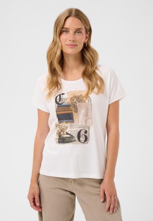 Kvinde med langt blond hår iført hvid T-shirt med vintage-stil grafik og beige bukser mod en ensartet lys baggrund.