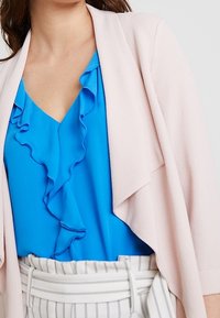 Ljusrosa kavaj över en blå volangblus med v-ringning, kombinerad med randiga byxor. Fokusera på mjuka texturer och lagerdesign.