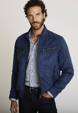 Homme barbu portant une veste en daim bleue par-dessus une chemise à motifs boutonnée, debout avec une main dans la poche, devant un fond uni.