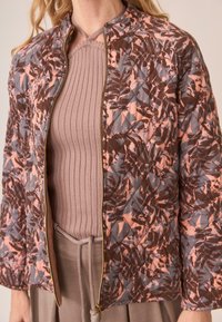 Mujer con una chaqueta acolchada con un patrón abstracto de hojas en rosa y marrón, sobre un top halter acanalado en taupe y pantalones a juego.