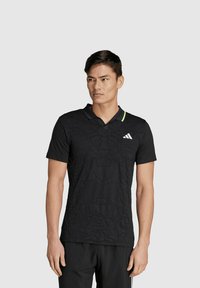 Zwart polo shirt met korte mouwen, voorzien van een textuurpatroon, een zwarte kraag met een groene accent, en een Adidas-logo op de linkerkant van de borst.