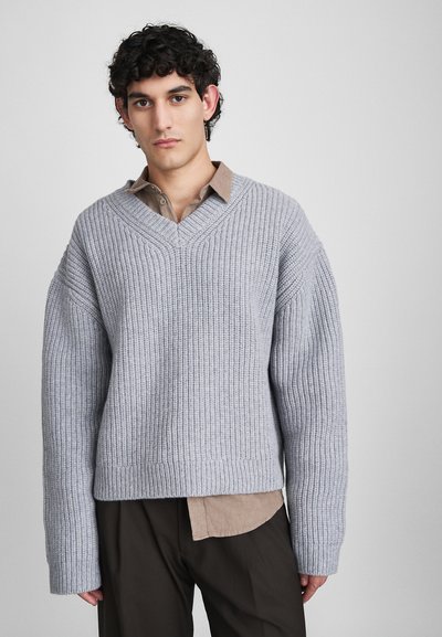 Un jeune homme avec des cheveux bouclés et foncés portant un pull en tricot côtelé gris ample par-dessus une chemise beige et un pantalon sombre, se tenant contre un fond uni.