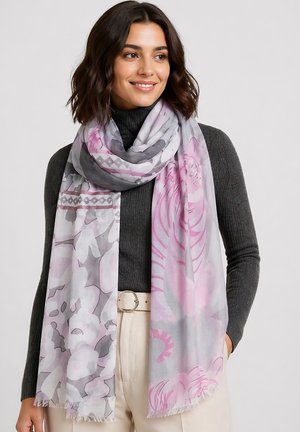 Frau, die einen hellrosa-grauen Blumenschal über einem dunkelgrauen Rollkragenpullover und cremefarbenen Hosen mit Gürtel trägt, lächelnd und leicht nach rechts schauend.