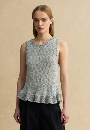 Frau trägt ärmelloses graues Strickoberteil mit Rüschensaum und schwarze Hose, steht vor einem schlichten beigen Hintergrund.