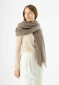 Écharpe en mélange de laine dans un doux dégradé de brun, avec des bords en franges, enroulée autour du cou et complétée par un haut beige clair et un pantalon blanc.
