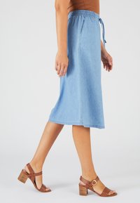 Jupe midi en denim bleu clair avec une taille élastique et un cordon de serrage noué. Associée à des sandales à talons strappy marron.