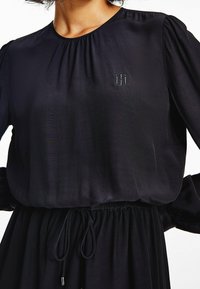 Schwarzes Kleid mit rundem Ausschnitt, Puffärmeln und tailliertem Bund. Verfügt über ein dezentes, gesticktes Logo auf der Brust. Glatte Stofftextur.