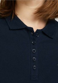 Marinblå pikétröja med krage, fem svarta knappar och ett texturerat stickat mönster.