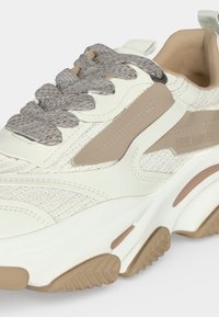 Gros plan d'une basket beige et blanche avec des lacets gris épais, une tige texturée, un col rembourré et une semelle épaisse avec des accents beiges.