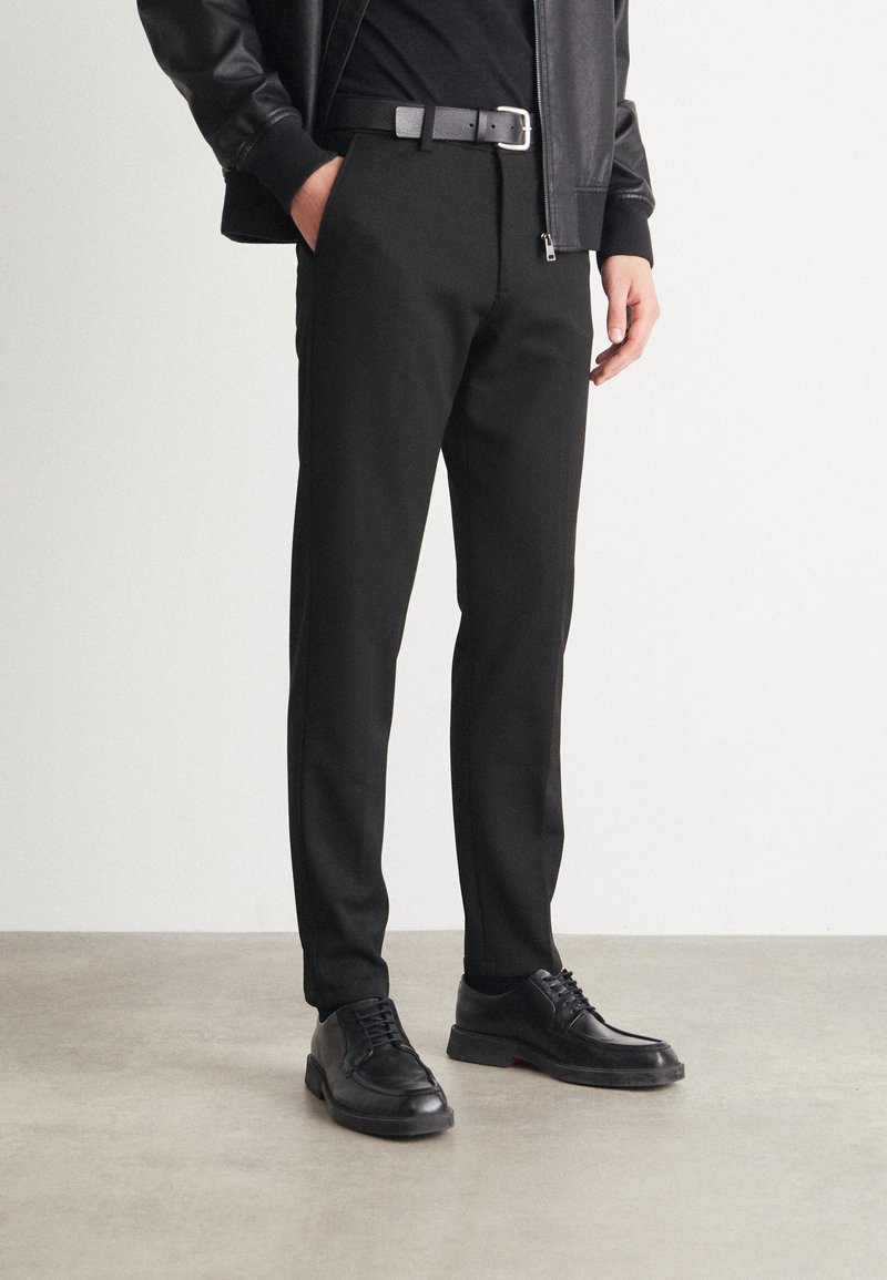 Pantalons noirs ajustés avec un design épuré, accompagnés d'une ceinture en cuir noir et de chaussures à lacets noires, sur un fond neutre.