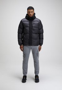 Giacca puffa nera con colletto alto e zip frontale, abbinata a pantaloni cargo grigi con tasche laterali e sneakers nere.