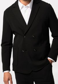 Blazer nero doppiopetto in tessuto texture, con rever a punta, due tasche frontali e bottoni neri. Camicia bianca visibile sotto.