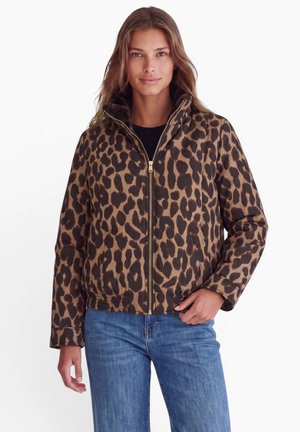 Femme portant une veste zippée imprimé léopard marron et noir avec un col montant et un jean bleu, debout sur fond blanc.