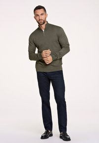 Olijfgroen gebreid pullover met een ritskraag, gecombineerd met marineblauwe broeken en zwarte schoenen. De stof lijkt soepel met een getailleerd ontwerp.