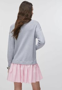 Pull gris à manches longues avec un ourlet arrondi, superposé sur une jupe plissée rose. Texture douce, coupe décontractée et design relax.