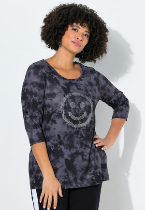 Kvinde med krøllet hår iført en sort og grå tie-dye langærmet top med en rhinsten-smiley, poserer med den ene hånd på hoften.