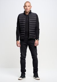 Crni puffer prsluk s vertikalnim šavovima, nošen preko crne majice dugih rukava. Slim-fit crne traperice i crne tenisice s bijelim potplatima.
