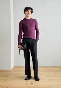 Maglione aderente viola a maniche lunghe abbinato a pantaloni neri su misura. Il modello tiene una borsa a tracolla nera. Texture liscia, design casual.