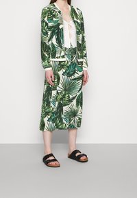 Ensemble en tissu doux composé d'une veste et d'une jupe à motif de feuilles vertes ; comprend un haut blanc avec découpe, complété par des sandales noires à deux lanières.