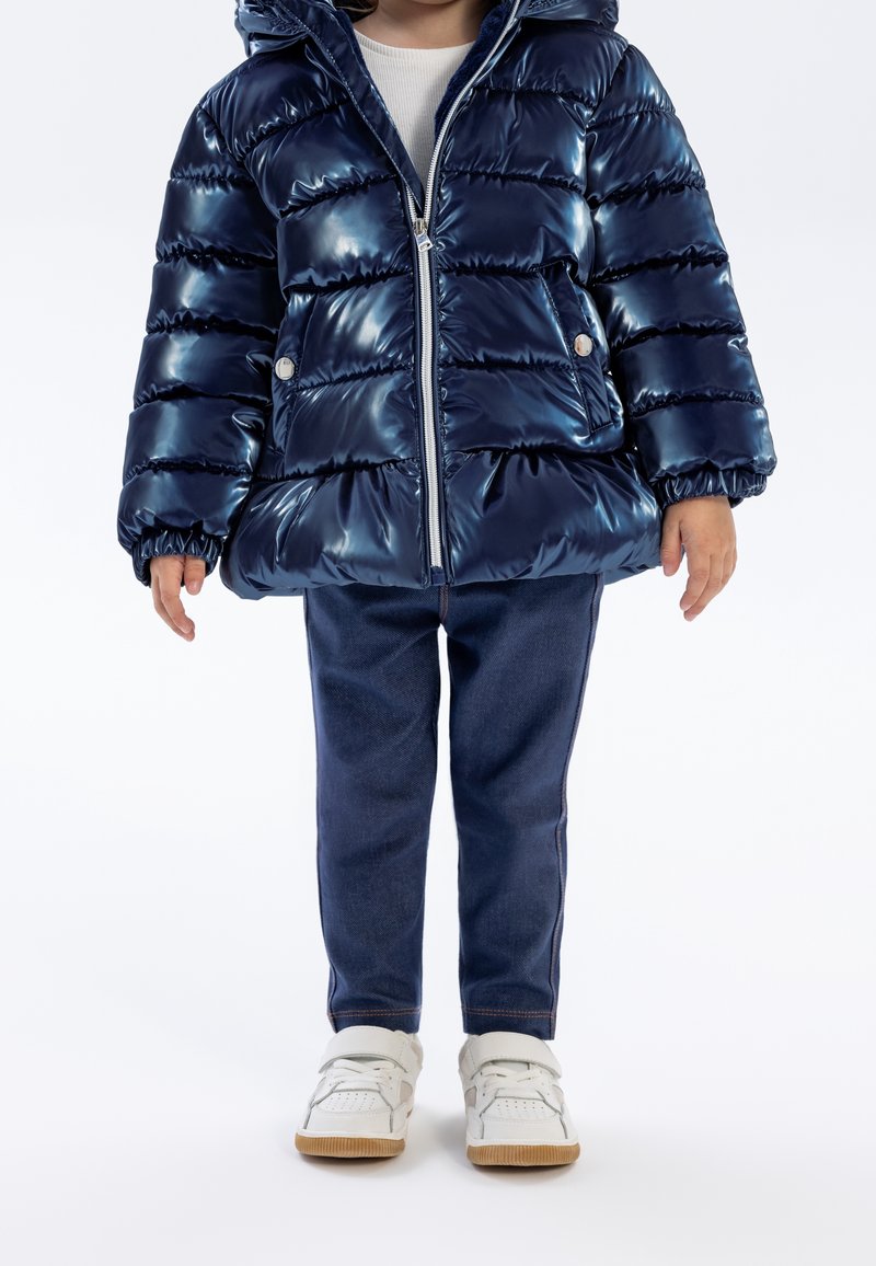 Enfant portant une doudoune bleu marine brillante, un pantalon bleu foncé, des baskets blanches et un haut blanc, debout devant un fond uni.