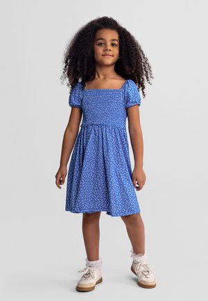 Dziewczynka o kręconych włosach w niebieskiej sukience w kwiaty z bufkami, białe skarpetki z falbanką i białe trampki, stojąca na tle szarego tła.