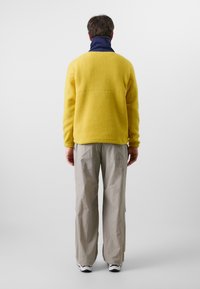 Geel fleece pullover met hoge hals, gecombineerd met lichtgrijze cargobroek. De gestructureerde stof en losse pasvorm zorgen voor comfort.