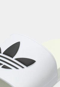 Λευκή σαγιονάρα με λευκή φάσα που φέρει το χαρακτηριστικό μαύρο λογότυπο τριφύλλι και τρεις μαύρες ρίγες, κατασκευασμένη από λείο, ανθεκτικό υλικό.