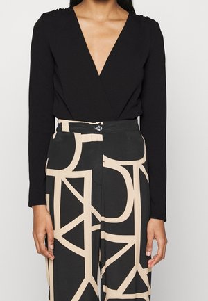 Vrouw draagt een zwart langarmig V-hals shirtje, ingestopt in een zwarte, hoge taille broek met beige abstracte geometrische patronen.