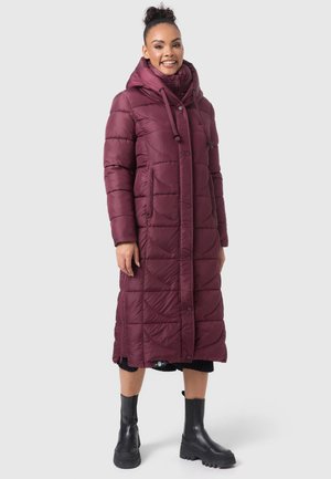 Lange bordeauxrode pufferjas met een quilted ontwerp, trekkoordcapuchon en zijzakken. Draagt in combinatie met kniehoge zwarte laarzen en een zwarte gebreide rok.