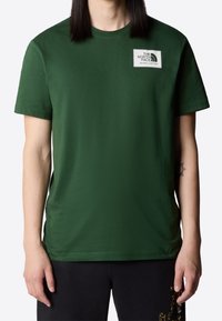 Osoba ubrana w ciemnozieloną koszulkę z krótkim rękawem z naszywką logo "The North Face" na piersi oraz czarne spodnie z żółtym wzorem.