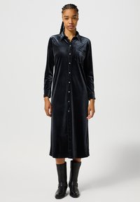 Abito camicia in velluto nero a maniche lunghe con chiusura a bottoni, taschino sul petto e una vestibilità comoda; abbinato a stivali neri fino al ginocchio.