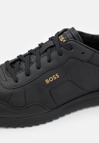 Černé sportovní boty s texturovanou tkaninou, hladkými doplňky a zlatým logem "BOSS". Mají kulaté tkaničky a pryžovou podrážku s vzorkem běhounu.