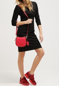 Röd crossbody-väska med guldfärgad hårdvara, rund form och justerbar axelrem, matchad med en svart klänning och röda sneakers.