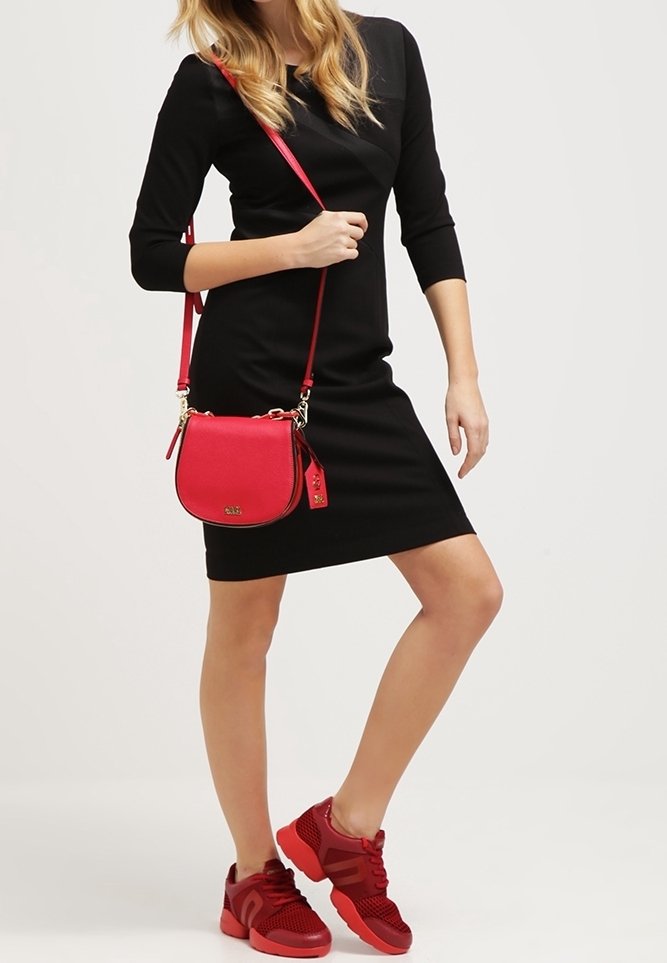 Röd crossbody-väska med guldfärgad hårdvara, rund form och justerbar axelrem, matchad med en svart klänning och röda sneakers.