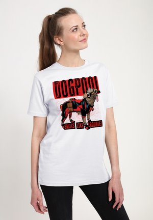 Młoda kobieta nosząca białą koszulkę z kreskówkowym psem w kostiumie superbohatera i napisem „Dogpool Smells Like Trouble.”